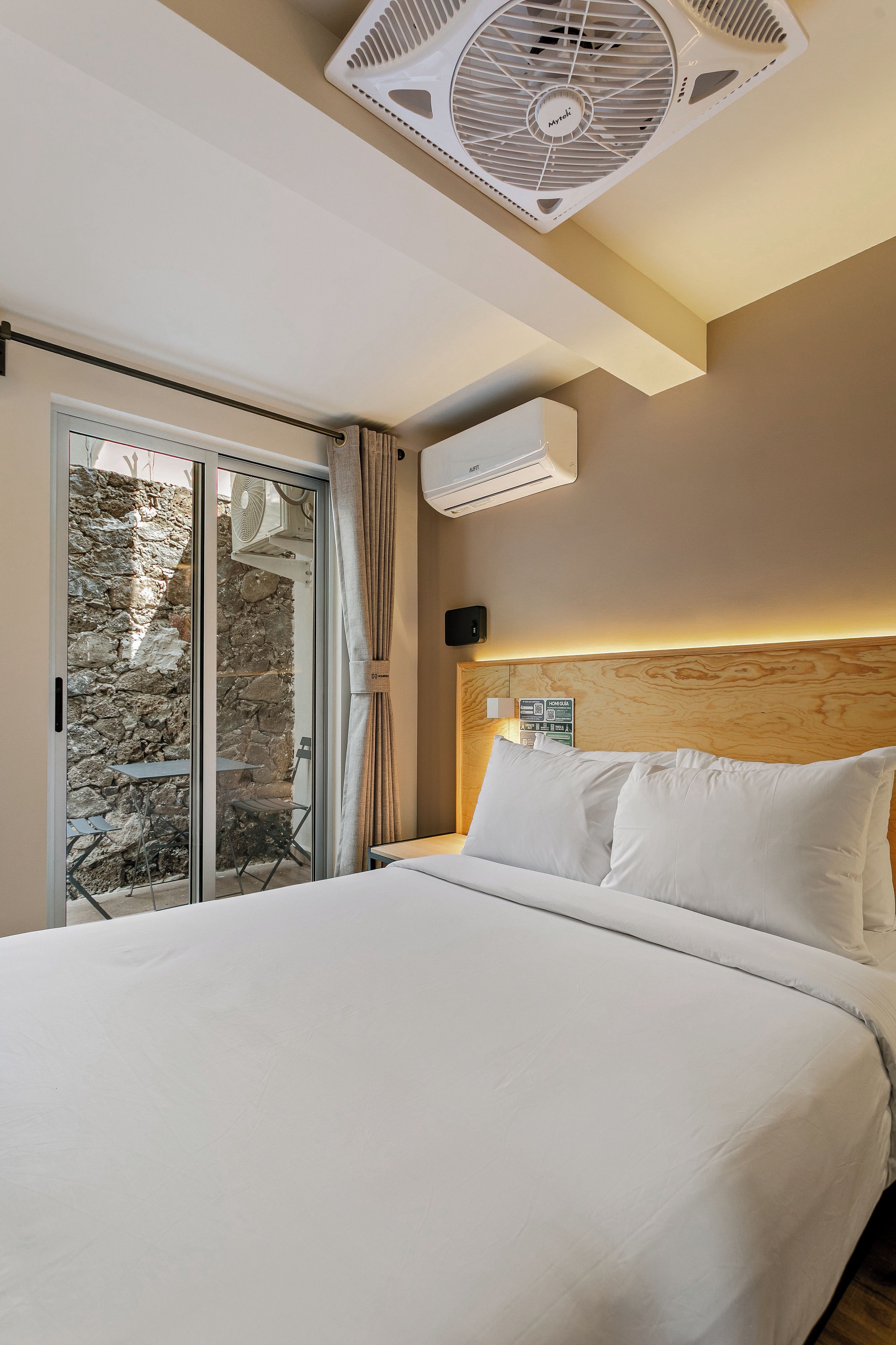 A hotel room with a bed, a ceiling fan, an air conditioning unit, a view through a glass door, and a wooden headboard.