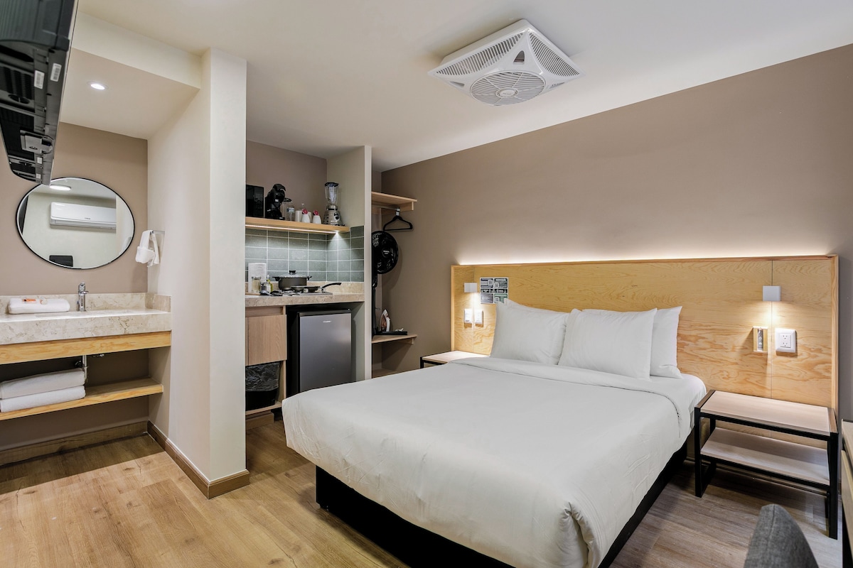 Habitación de hotel con cama, mesita de noche, espejo, kitchenette pequeño y baño.