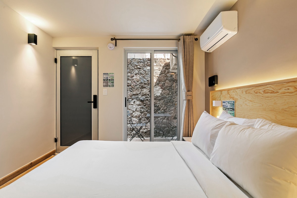 Habitación de hotel con cama, puerta, ventana con vista a árboles y un equipo de aire acondicionado.