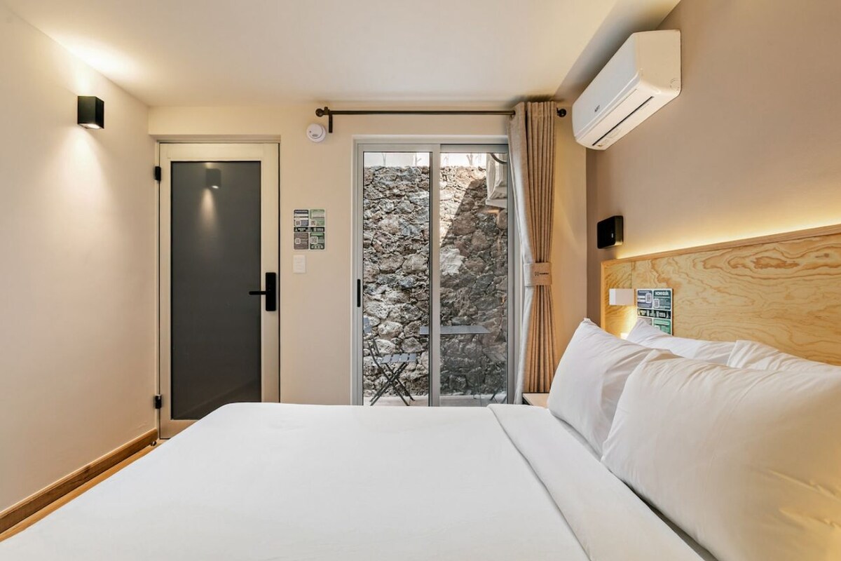 Habitación de hotel con una cama, una puerta, un equipo de aire acondicionado, una ventana con vista a un paisaje rocoso y una lámpara fijada a la pared.