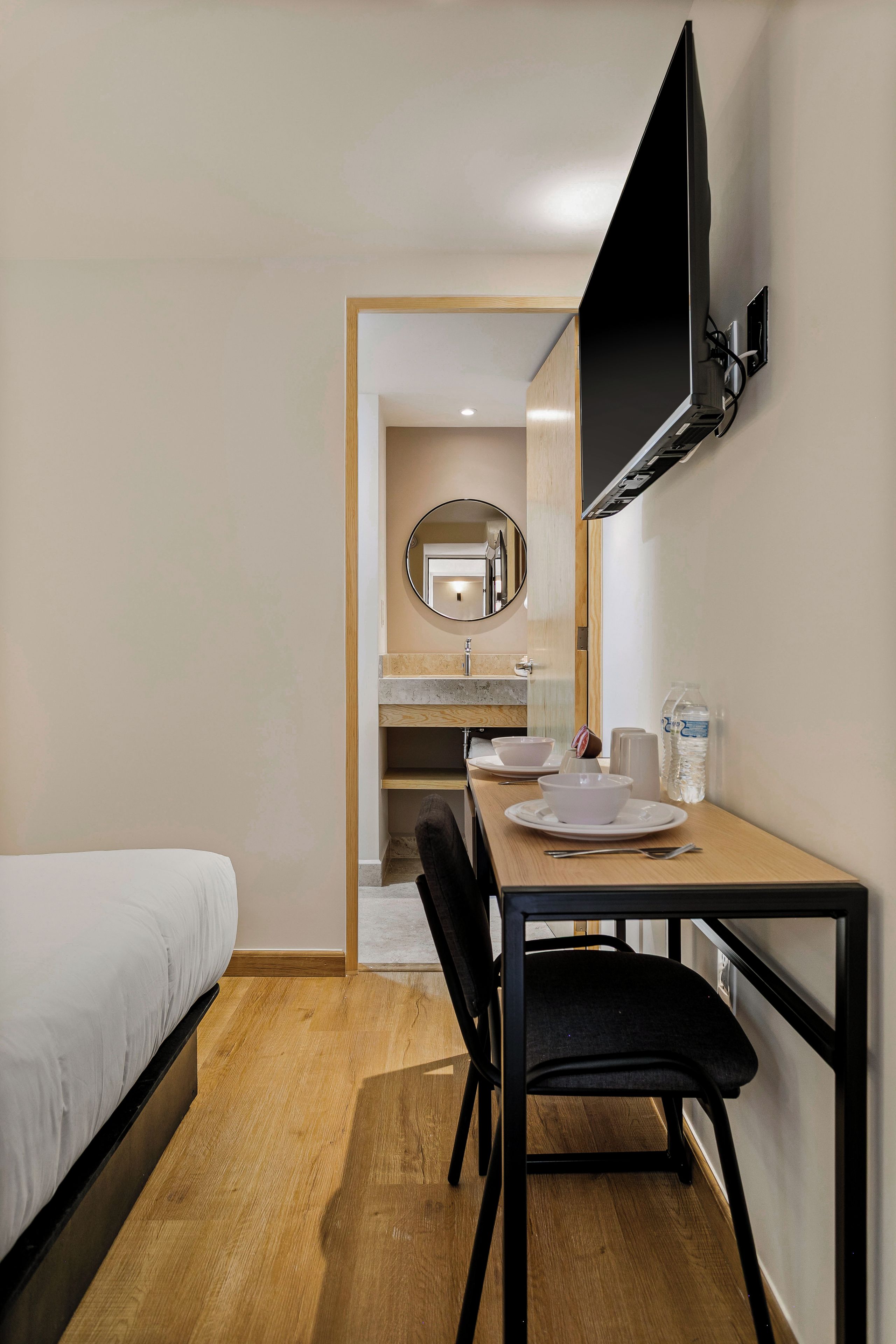 Une chambre d’hôtel avec un lit, un bureau et une chaise, un téléviseur fixé au mur, un miroir et une salle de bain visible à travers une porte ouverte.