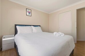 A neatly made bed with white linens, a bedside table with a lamp, and a door to the right. - The Wattle House - Your Home Away from Home (Wattle Grove)