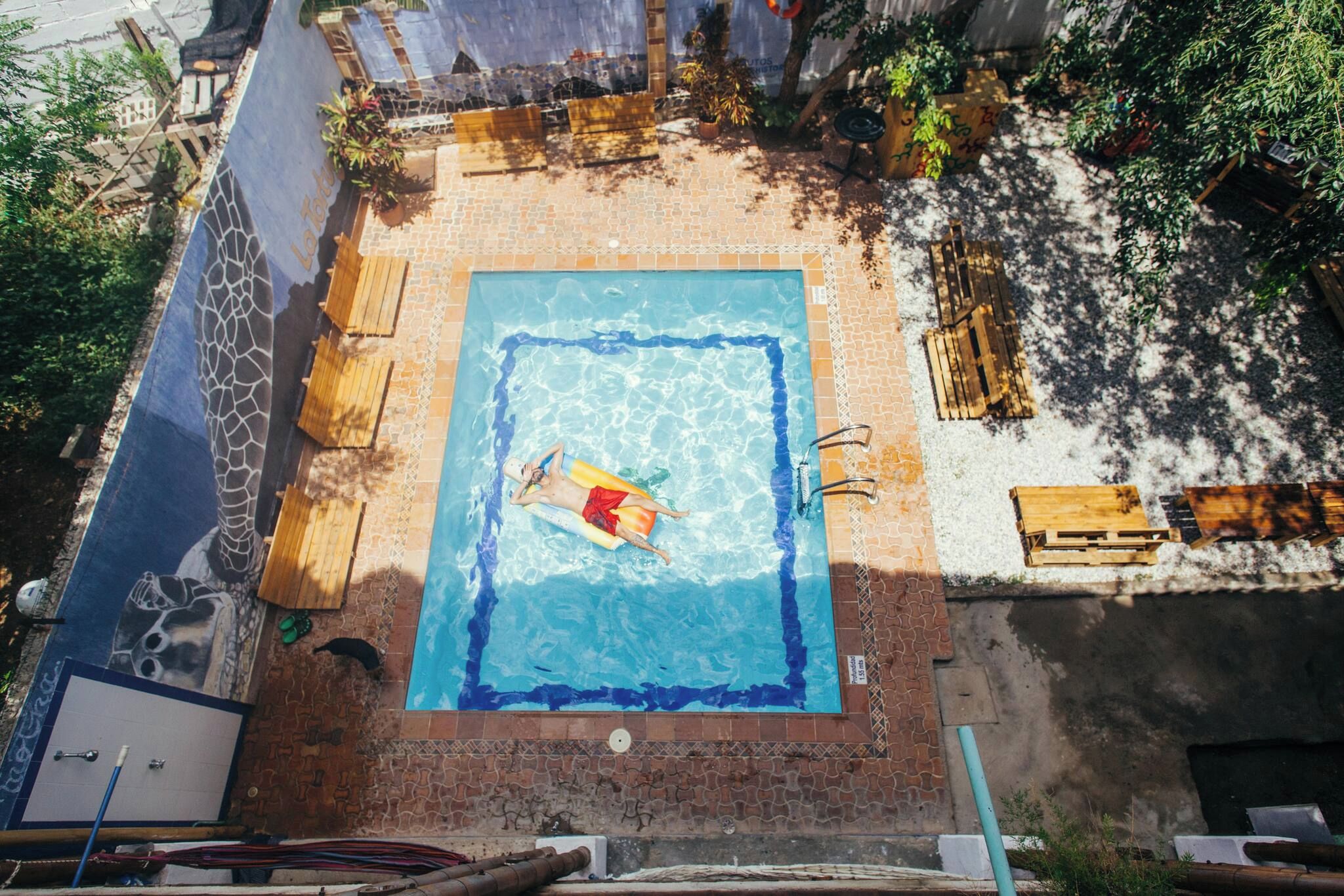 A rooftop swimming pool with a person in the water, surrounded by a brick patio and wooden furniture.
