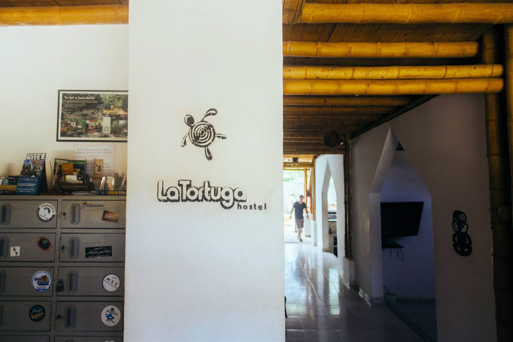 A hostel interior with a central pillar displaying the hostel's logo, a wall-mounted sign, and a corridor leading to another room.