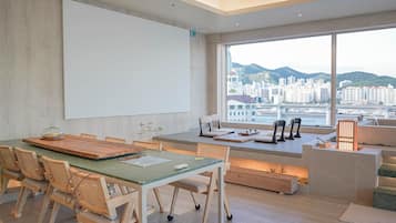 Una sala de reuniones moderna con una mesa grande, sillas, una pantalla de proyector y vistas de un paisaje urbano a través de amplios ventanales.