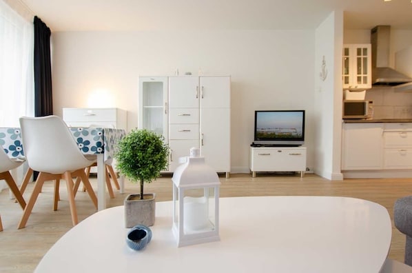 Ett modernt vardagsrum med ett vitt matbord, stolar, en liten krukväxt, en vit lykta och en tv som står på ett tv-bord.
