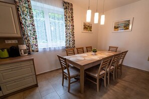 Un comedor con una mesa y sillas de madera, una ventana con cortinas y una zona de cocina con armarios y electrodomésticos.