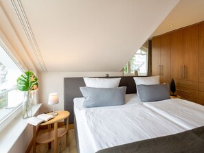 A bedroom with a large bed, a bedside table with a lamp, a window with a view, and a wooden wardrobe. - Penthouse Villa Gudrun With Private Sauna (Binz)