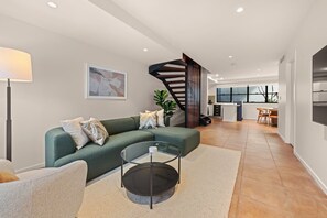 A modern living room with a green sofa, a glass coffee table, and a staircase leading to an open kitchen area. - Spacious 3-Level Windsor Townhouse | 2 Carparks (Brisbane)
