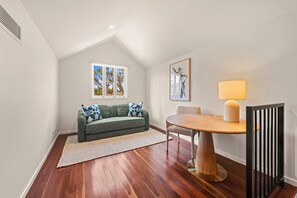 A room with a wooden floor, a green sofa with patterned cushions, a round wooden table, a lamp, and a window with a view of trees. - Spacious 3-Level Windsor Townhouse | 2 Carparks (Brisbane)
