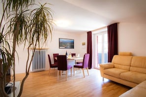 A living room with a sofa, a dining table with chairs, a radiator, and a large window with curtains. - Melanie's Guesthouse (Kaltern)