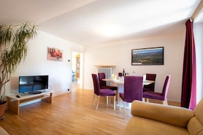 Un salon avec un téléviseur, un coin repas avec des chaises violettes et un canapé.