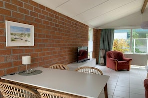 A room with a brick wall, a dining table with chairs, a television, a red armchair, and a window with curtains. - Holiday home with 1 bedroom (Burgerbrug)