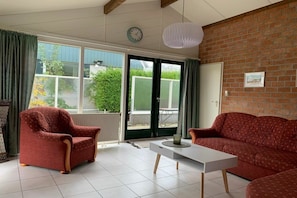 A living room with a sofa, armchair, and a coffee table. There is a window with curtains, a door, and a brick wall. - Holiday home with 1 bedroom (Burgerbrug)