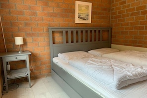 Een kamer met een bakstenen muur, een bed met witte beddengoed, een nachtkastje met een lamp, en een ingelijst kunstwerk aan de muur.