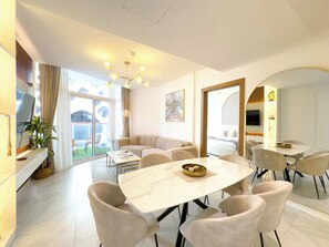 A modern living room with a sofa, a dining table with chairs, a chandelier, and a large window. - Rentico | 1BR Burj Khalifa & Canal View Balcony (Dubai)