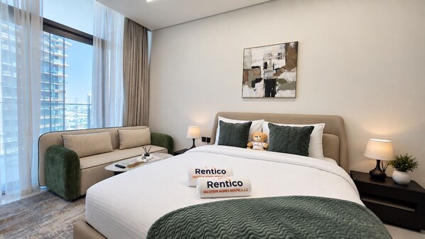 Habitación de hotel con cama, sofá, mesita de noche, lámpara y ventana con cortinas.