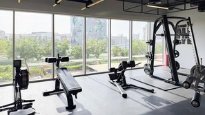 Een sportschool met diverse fitnessapparatuur, grote ramen en uitzicht op gebouwen en bomen.