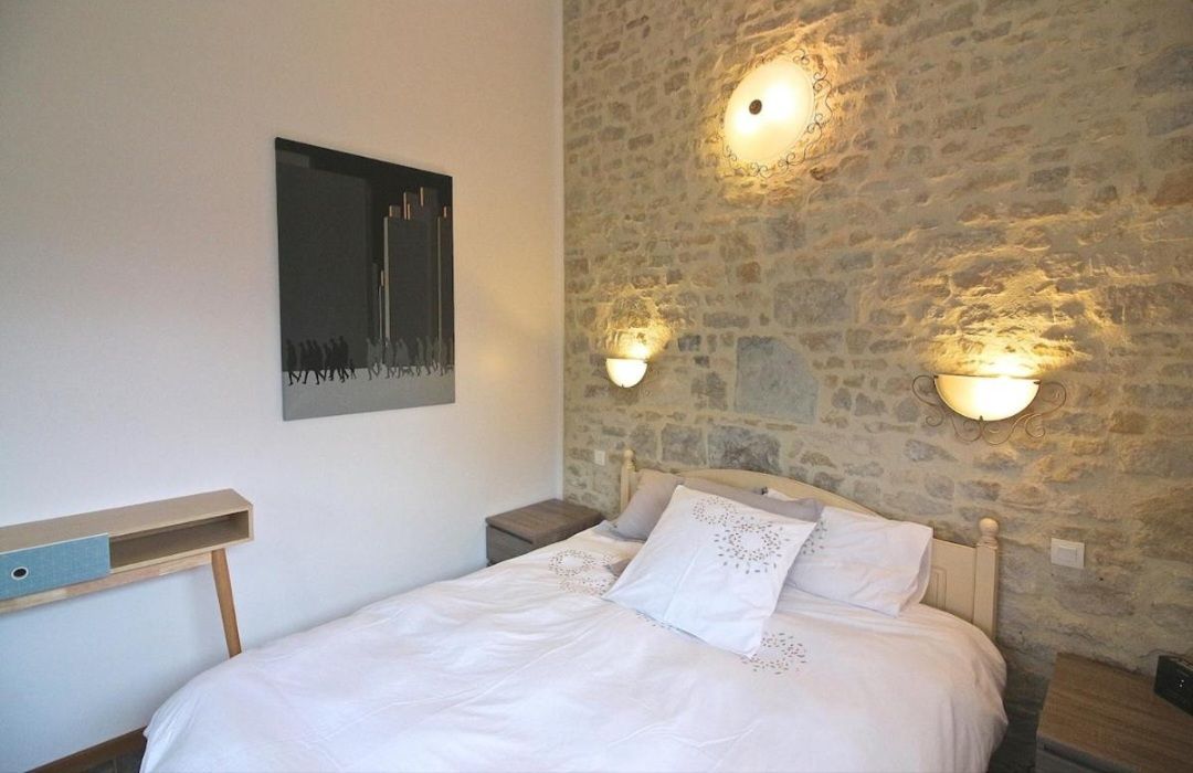 Un dormitorio con pared de piedra, una cama con ropa de cama blanca, dos lámparas de pared y una mesita de noche pequeña.