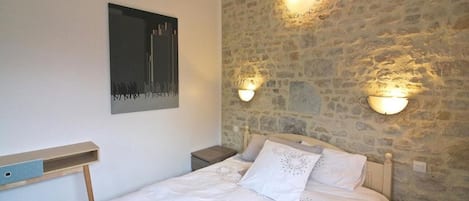 A bedroom with a stone wall, a bed with white bedding, two wall-mounted light fixtures, and a small bedside table.