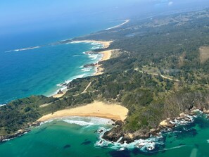 Eine Luftaufnahme einer Küstenregion mit Strand, bewaldeten Hügeln und freiem Blick auf den Ozean.