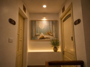 Un couloir avec des portes portant les numéros 5A et 5B, un tableau représentant une fenêtre avec un livre, et une plante en pot.