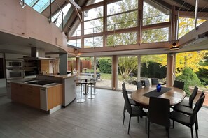 A modern kitchen with a central island, dining area with chairs, and large windows offering a view of the outdoors.