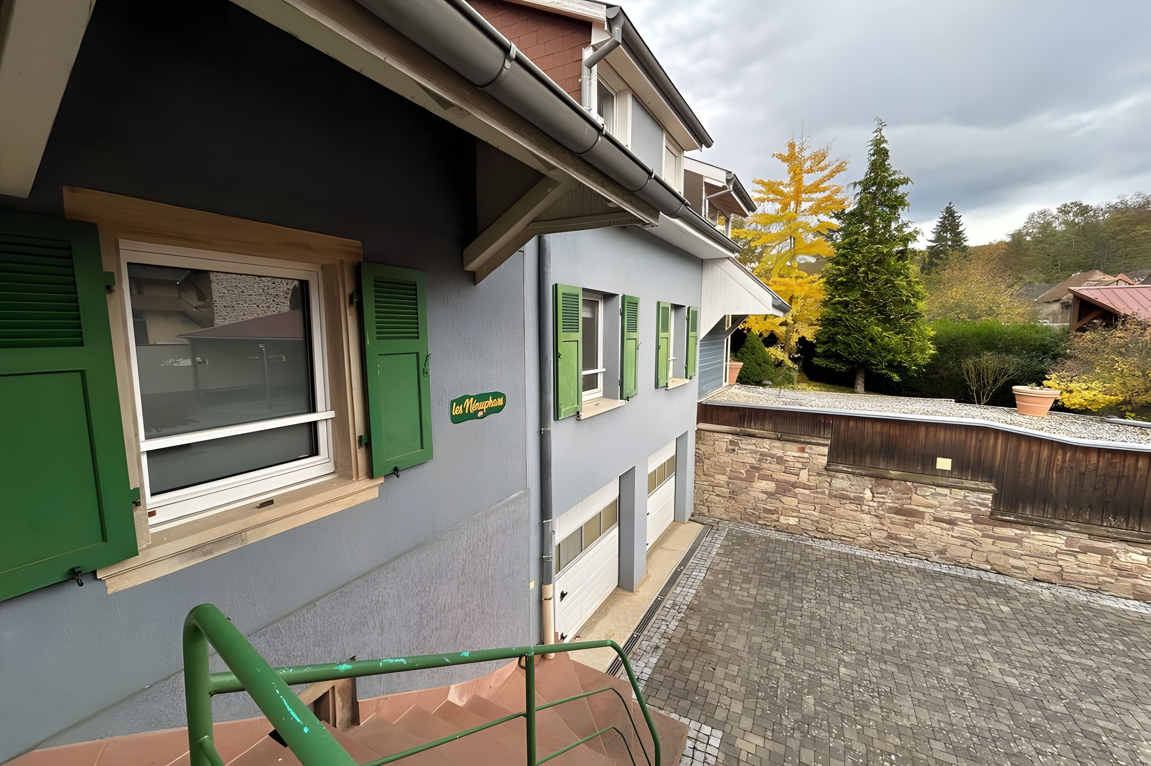 Un edificio residencial con persianas verdes, un muro de piedra y una zona pavimentada con barandilla metálica.