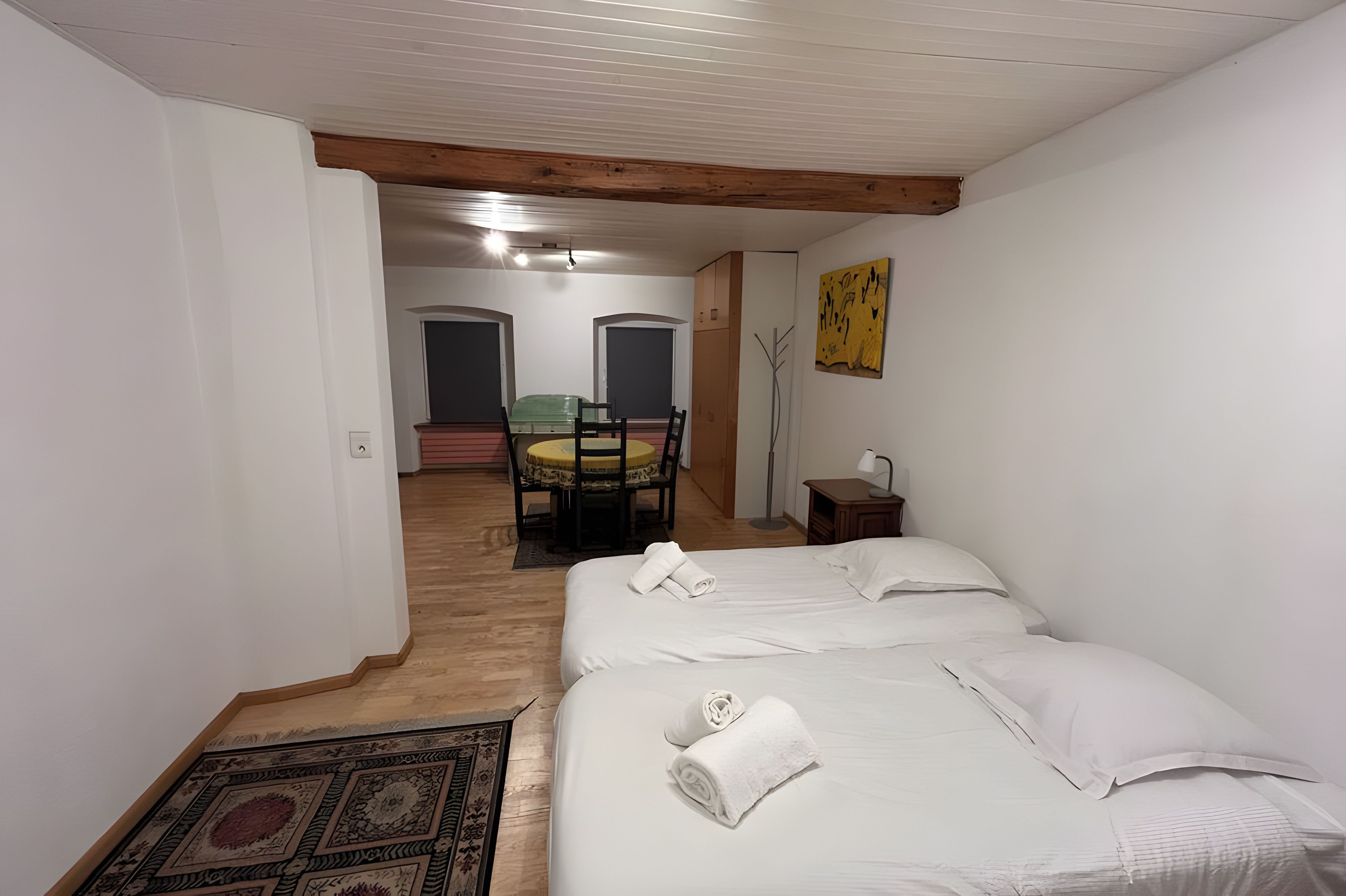 Una habitación con una cama, una mesita, una silla y una lámpara. Se ve una puerta que da a otra habitación.