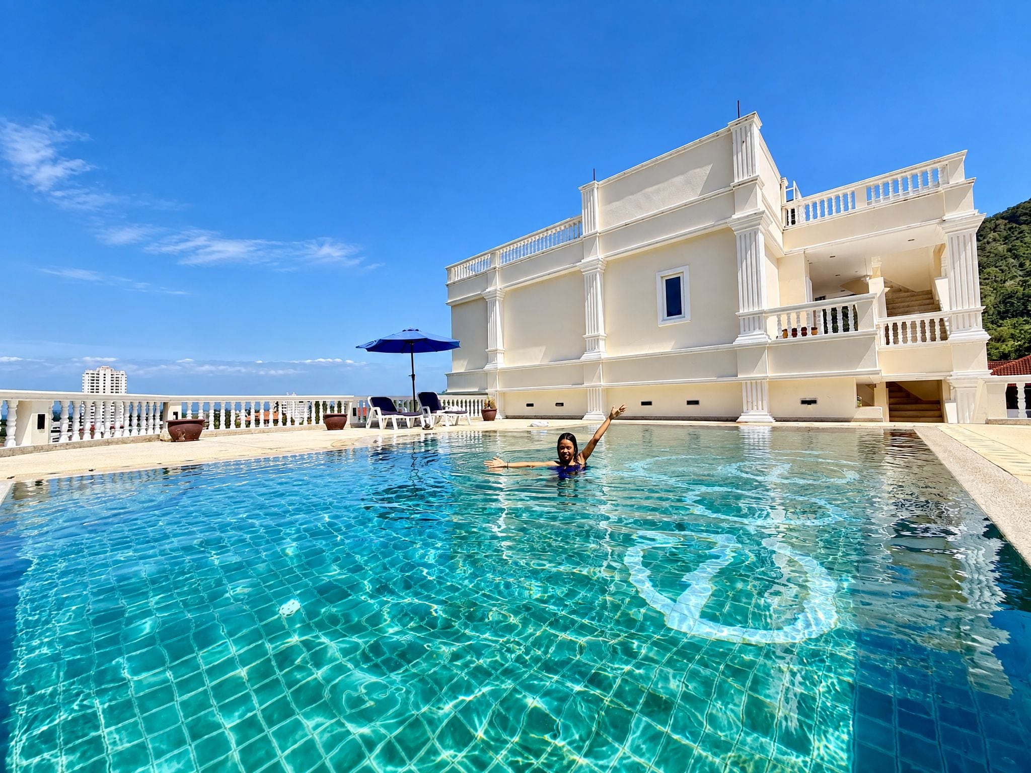 Piscina com uma pessoa na água, um prédio branco com varandas e céu azul límpido.