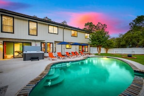 A pool area with lounge chairs and umbrellas, a barbecue grill, and a building with multiple windows. - Pool Haven | Hot Tub Game Room Fire Pit Mini Golf (San Antonio)