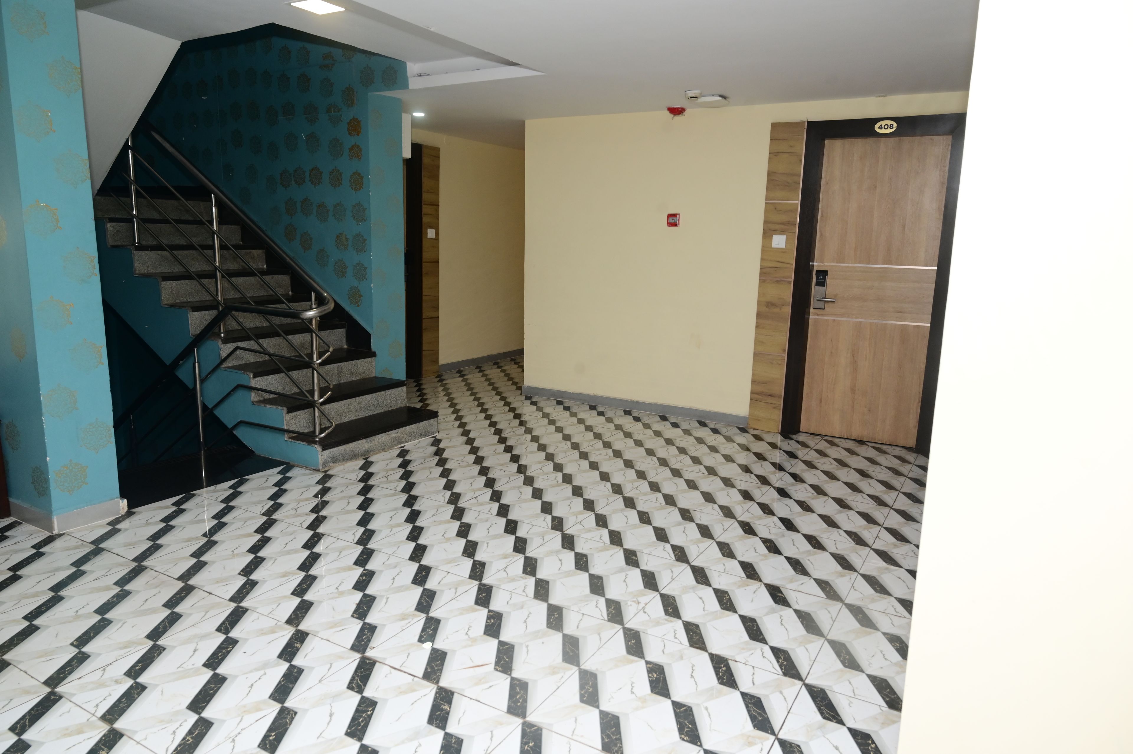 Un vestíbulo de hotel con piso a cuadros, una pared azul con diseño geométrico, una escalera con barandas de metal y una puerta con placa de número.