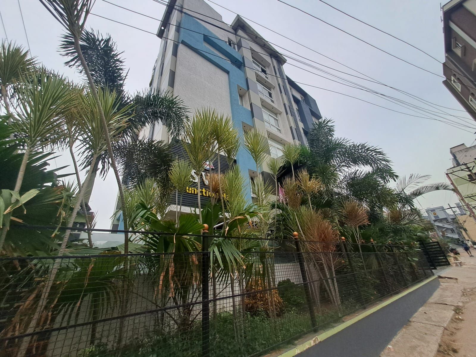 A multi-story building with a blue and white facade, surrounded by a fence and various tropical plants.