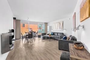 A modern living room with a grey sofa, a dining area with a table and chairs, and a large window offering a view of a colorful building. - Spacious holiday apartment with luxury features and covered terrace near the Bal (Lübeck)