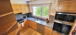 A modern kitchen with wooden cabinets, a granite countertop, a built-in oven, and a window with a view of greenery. - Cozy vacation rental for families with mountain views, midday sun, a pool, and a sauna (Davos)