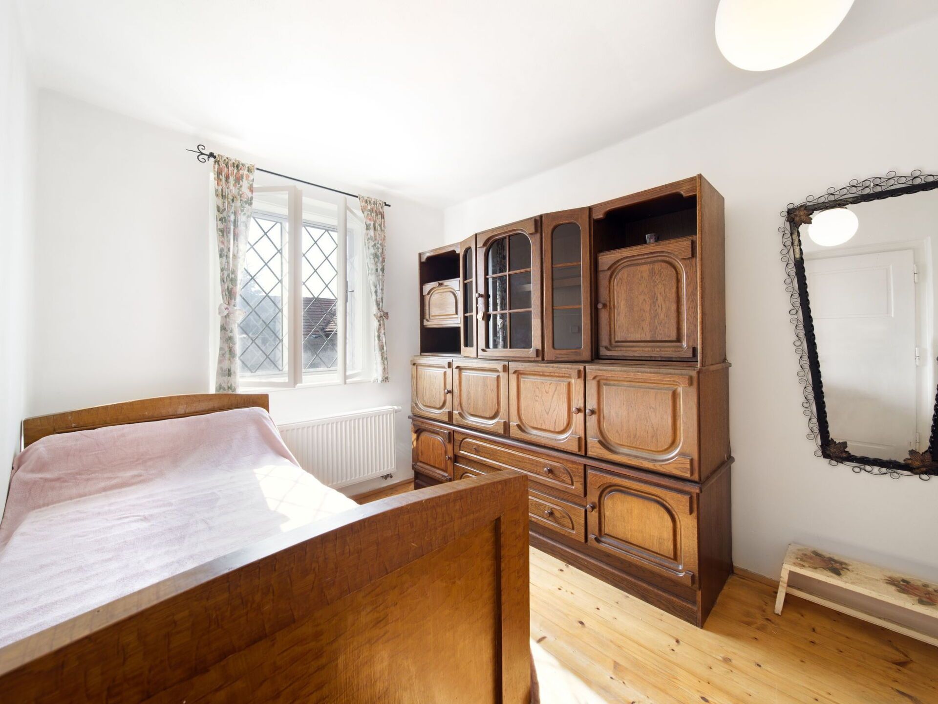 Une chambre avec un lit en bois, une grande armoire en bois, une fenêtre avec des rideaux et un miroir au mur.