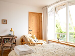 Ein gemütliches Schlafzimmer mit einem großen Fenster, einer Holztür, einem Sessel mit Decke und einem Nachttisch mit Lampe.