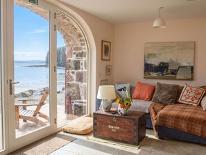 A coastal living room with a stone wall, a sofa, a coffee table, and a view of the sea. - Beautiful private villa for 6 people with WIFI and patio (Isle Of Skye)
