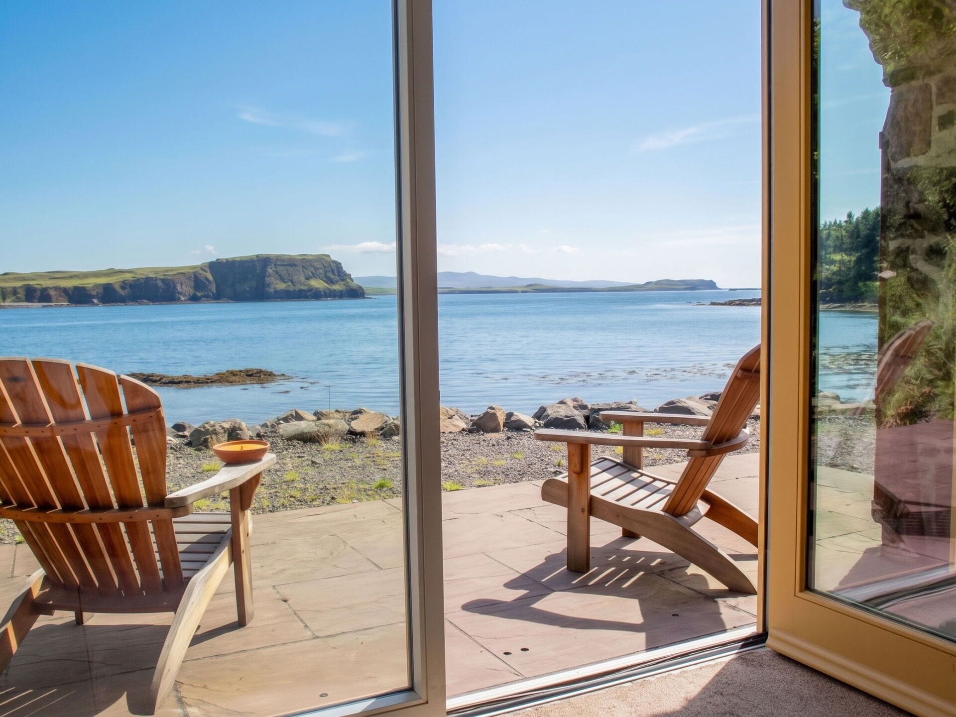 A view through a glass door to a patio with two wooden chairs and a table, overlooking a body of water with a rocky shoreline and distant landforms.