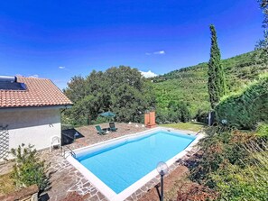 En swimmingpool med ett kaklat poolområde, en loungehörna med stolar och utsikt över en sluttning.