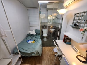 Ein kleines Hotelzimmer mit Bett, Schreibtisch, Stuhl, Nachttisch, Nachttischlampe, einer Uhr, einem Spiegel und einer Duschkabine.