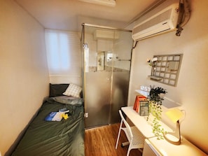 Ein kleines Zimmer mit Bett, Schreibtisch, Dusche und einer Wand-Klimaanlage.
