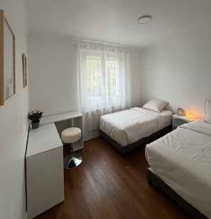 Quarto com duas camas, uma escrivaninha, uma cadeira e janela com cortinas.