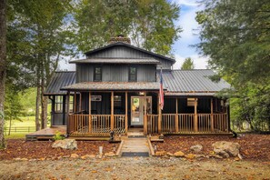 A wooden cabin with a porch, a flag, and a stone chimney. - Pond in the Pines | Aska Farm Stay + Fishing (Blue Ridge)