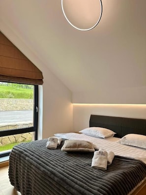 Quarto de hotel com uma cama grande, cabeceira e vista para a estrada e vegetação lá fora.