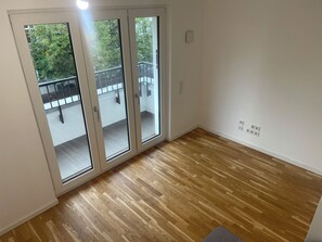Ein Innenraum mit Holzboden, einer Schiebeglasdoor zu einem Balkon im Freien und einer weißen Wand mit Steckdosen.