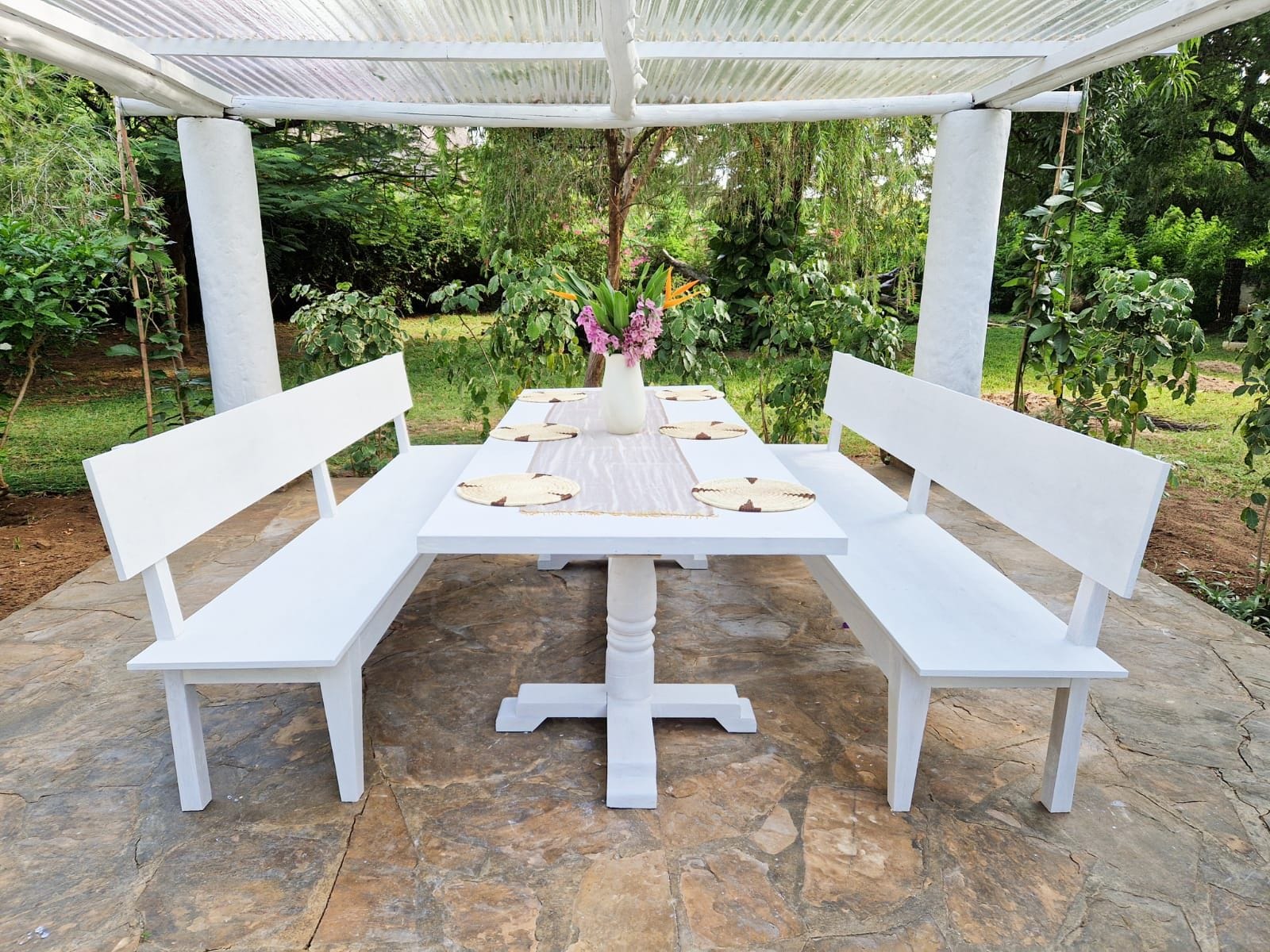 Ein weißer Tisch im Freien mit einer Bank an jeder Seite, auf einer Steinterrasse mit Garten im Hintergrund.