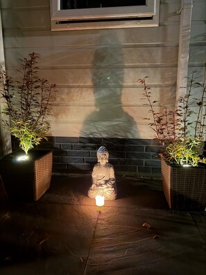 A statue of a seated figure with a light source in front, flanked by two potted plants, against a wall with a window above. - Chalet Lebensfreude – A Getaway by the Water (Lathum)