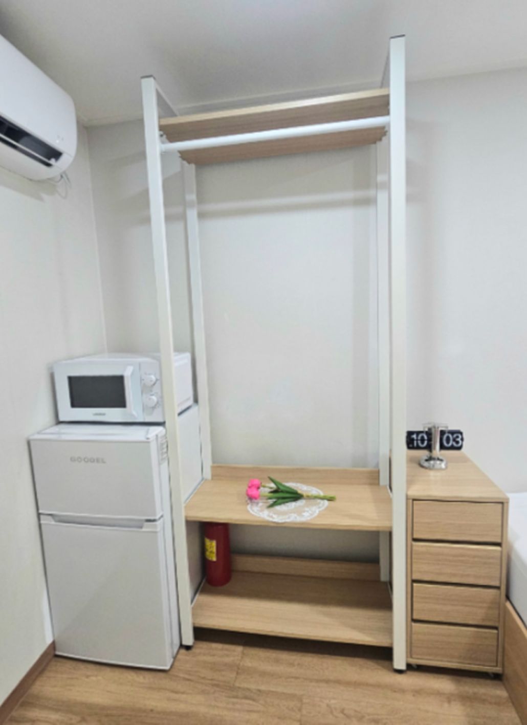 A compact room with a bed, a small kitchenette with a microwave and refrigerator, a bedside table with a clock, and a wooden bench with a flower.