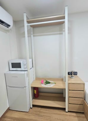 Une chambre compacte comprenant un lit, une petite kitchenette équipée d’un four à micro-ondes et d’un réfrigérateur, une table de chevet avec une horloge, et un banc en bois agrémenté d’une fleur.